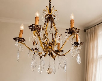 May include: A gold-toned metal chandelier featuring five candle-style light fixtures with beige sleeves. The frame is decorated with ornate floral metalwork and numerous clear, faceted crystal teardrop pendants hanging from the curved arms.