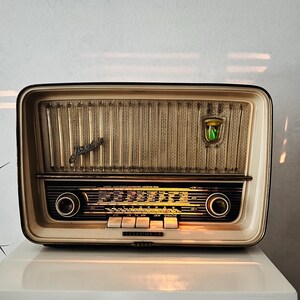 May include: A cream-coloured Telefunken radio with a rectangular shape, featuring a textured beige speaker grille, a glowing green indicator light, and a black control panel with yellow text, knobs, and four white push-button switches.