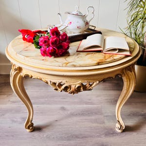 May include: A round coffee table with a white marble top and ornate gold painted legs. The table has a bouquet of red roses and a book on top.