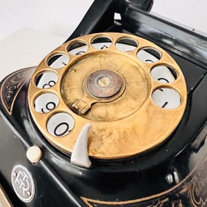 May include: Close-up of a vintage black rotary dial telephone with a gold-colored dial and a silver finger stop. The phone has ornate gold detailing and a white button. A classic communication device.