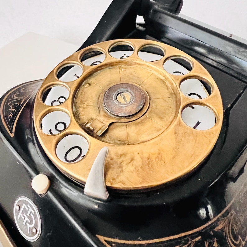 May include: Close-up of a vintage black rotary dial telephone with a gold-colored dial and a silver finger stop. The phone has ornate gold detailing and a white button. A classic communication device.