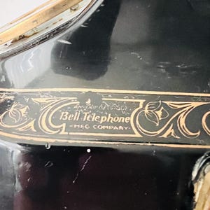 May include: Close-up of a black antique telephone with gold-colored decorative accents. The text "Bell Telephone - MFG COMPANY" is visible in a gold-colored font. The phone has a glossy finish.