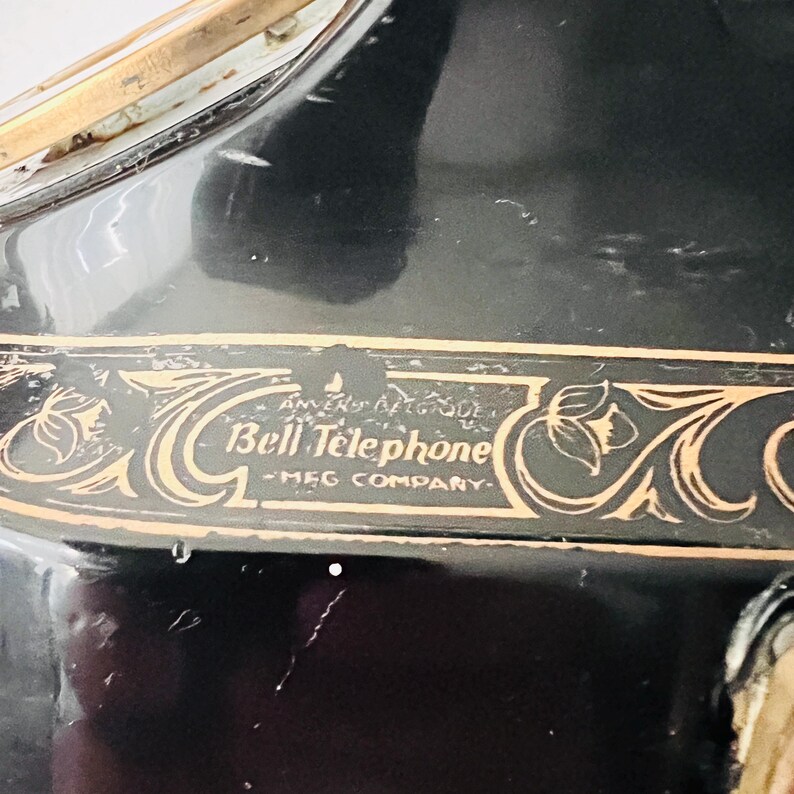 May include: Close-up of a black antique telephone with gold-colored decorative accents. The text "Bell Telephone - MFG COMPANY" is visible in a gold-colored font. The phone has a glossy finish.