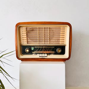 Könnte beinhalten: Ein Vintage-Radio von Telefunken mit einem Gehäuse aus poliertem Holz und einer cremefarbenen Frontblende. Das Gerät verfügt über horizontale, goldfarbene Lautsprecherlamellen, eine zentrale Skala mit schwarzem Hintergrund, zwei Drehknöpfe und den Schriftzug Gavotte.