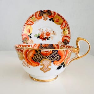 Op de afbeelding: Een vintage theekopje en schotelset met een bloemenpatroon in tinten oranje, rood, blauw en goud. Het kopje en de schotel hebben een gegolfde rand en een gouden rand.