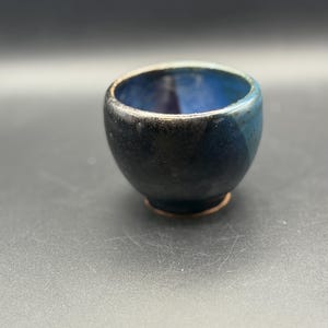 May include: A small, rounded ceramic cup featuring a deep blue interior glaze that transitions into a dark, mottled black and navy exterior. The rim is slightly lighter in tone, and the base reveals a thin, unglazed copper-colored ring.