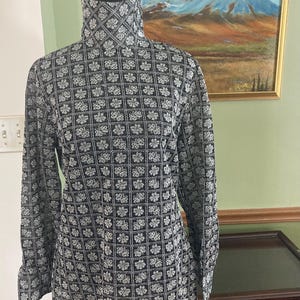 May include: A black and white patterned long-sleeve shirt with a high collar. The shirt features a repeating floral design in a grid pattern. The garment is displayed on a mannequin. The background includes a painting of a mountain landscape.