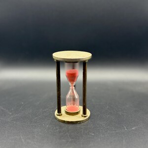 May include: A small hourglass featuring a metallic gold-toned frame with a circular top and base supported by three vertical pillars. The central glass bulb contains fine red sand, with a portion currently flowing from the top to the bottom chamber.