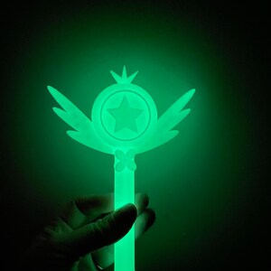 May include: A green glow-in-the-dark wand with a star and wings. The wand is made of plastic and has a smooth surface.