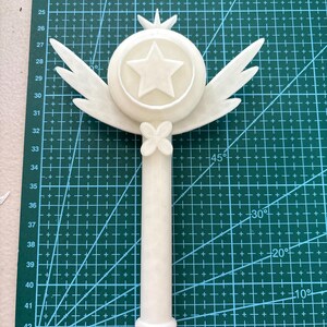 May include: White plastic wand with a star and wings on top. The wand is on a green cutting mat with markings in inches.