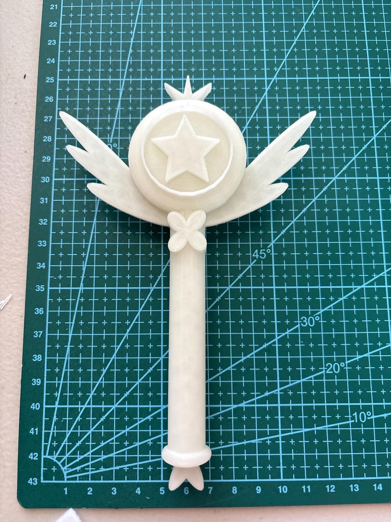May include: White plastic wand with a star and wings on top. The wand is on a green cutting mat with markings in inches.