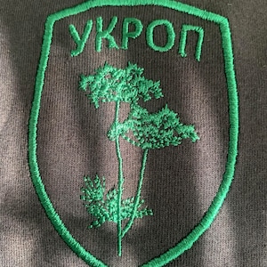 Peut inclure: Patch brodé vert en forme de bouclier, avec une plante d'aneth stylisée et le mot "УКРОП" en lettres cyrilliques.