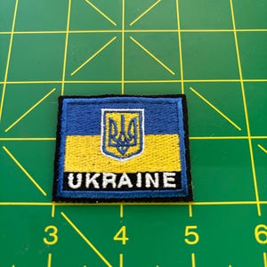 May include: A blue and yellow embroidered patch with the Ukrainian coat of arms and the word "UKRAINE" in white lettering.