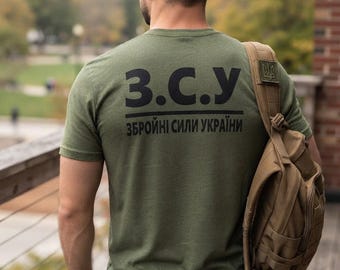 May include: An olive green short-sleeve t-shirt featuring the black text Z.C.Y and ZBROINI SYLY UKRAINY printed on the back. The shirt is worn with a tan tactical backpack that has a small patch with a trident emblem on the shoulder strap.
