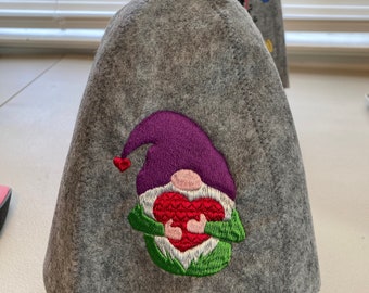 May include: Gray felt teapot cozy with a gnome holding a red heart embroidered on the front. The gnome has a purple hat and green sleeves.