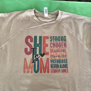 May include: A beige sweatshirt with a colorful graphic print that reads "She is Mom" with a heart symbol. The graphic also includes the words "Strong, Chosen, Beautiful, Capable, Victorious, Never Alone, Greatly Loved".