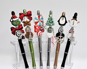 May include: A collection of decorative pens with Christmas-themed toppers, including a Grinch, gingerbread man, and snowman. The pens have black, green, silver, and brown barrels, displayed in a clear acrylic holder. Each pen features a unique charm.