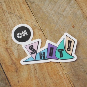 May include: A sticker with the words "OH SHIT" in a colorful, geometric design. The sticker features a black circle with "OH" in white, and the word "SHIT" in various shapes and colors, including pink, blue, purple, and teal.