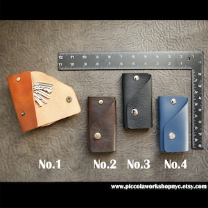 May include: Four leather keychains in different colors and styles. The keychains are numbered 1 through 4. The keychains are on a brown textured surface. The text "www.piccolaworkshopnyc.etsy.com" is at the bottom of the image.