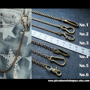 May include: A selection of six brass-toned wallet chains with various clasp styles. The chains are displayed against a dark background with a ruler for scale. One chain is attached to a pair of denim jeans with star embellishments. The website address is visible at the bottom.
