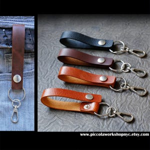 May include: Leather keychains in black, brown, and tan. Each keychain features a leather strap with a metal snap and a silver-colored clasp. One keychain is attached to a pair of blue jeans.