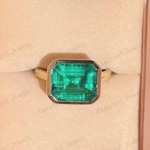 Könnte beinhalten: Ein goldener Ring mit einem großen, rechteckigen, smaragdgrünen Edelstein. Der Ring wird in einer beigen Schmuckschatulle mit rosa Satinfutter präsentiert. Die Schatulle wird geöffnet gehalten.