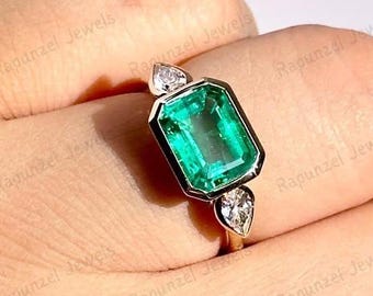 May include: A gold ring featuring an emerald-cut, vibrant green gemstone. Two smaller, pear-shaped clear gemstones flank the main stone. The ring is set on a gold band, showcasing a classic and elegant design.