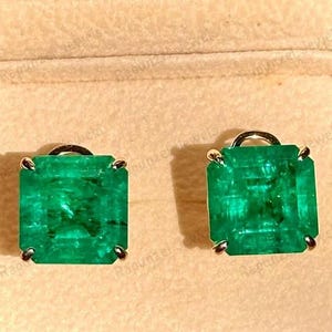 Puede incluir: Un par de pendientes de esmeralda de talla esmeralda engastados en oro, expuestos dentro de un joyero de madera con interior beige. Los pendientes son de color verde vibrante y tienen forma cuadrada.