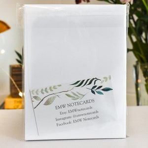 K&ouml;nnte beinhalten: Ein Paket wei&szlig;er Notizkarten, verpackt in durchsichtigem Zellophan. Eine kleine Karte mit einem Aquarelldesign von Bl&auml;ttern und dem Text "EMW NOTECARDS" ist sichtbar. Der Hintergrund umfasst Pinsel und eine Blumenvase.
