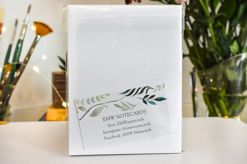 K&ouml;nnte beinhalten: Ein Paket wei&szlig;er Notizkarten, verpackt in durchsichtigem Zellophan. Eine kleine Karte mit einem Aquarelldesign von Bl&auml;ttern und dem Text "EMW NOTECARDS" ist sichtbar. Der Hintergrund umfasst Pinsel und eine Blumenvase.