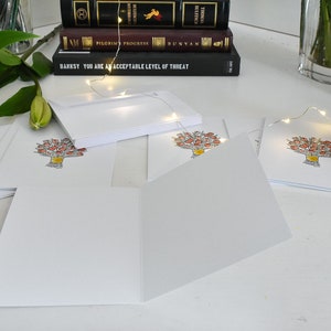 K&ouml;nnte beinhalten: Leere wei&szlig;e Gru&szlig;karten mit floralem Design. Die Karten sind offen und gestapelt, mit einer Aquarellillustration eines Blumenstrau&szlig;es in einer gelben Vase. B&uuml;cher und Blumen sind im Hintergrund.