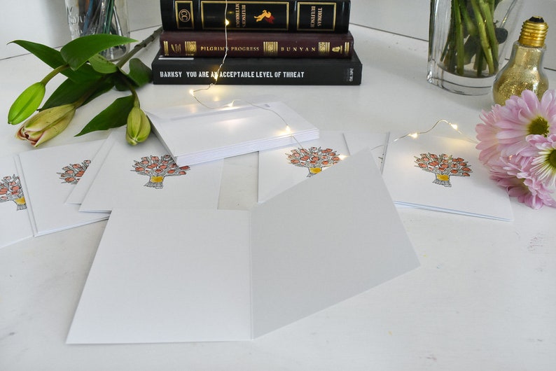 K&ouml;nnte beinhalten: Leere wei&szlig;e Gru&szlig;karten mit floralem Design. Die Karten sind offen und gestapelt, mit einer Aquarellillustration eines Blumenstrau&szlig;es in einer gelben Vase. B&uuml;cher und Blumen sind im Hintergrund.