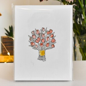 K&ouml;nnte beinhalten: Eine wei&szlig;e Gru&szlig;karte mit einer Aquarellillustration eines Bouquets aus rosa Tulpen, die mit einem gelben Band gebunden sind. Die Karte ist in einer transparenten Plastikh&uuml;lle versiegelt, mit Pinseln und Blumen im Hintergrund.