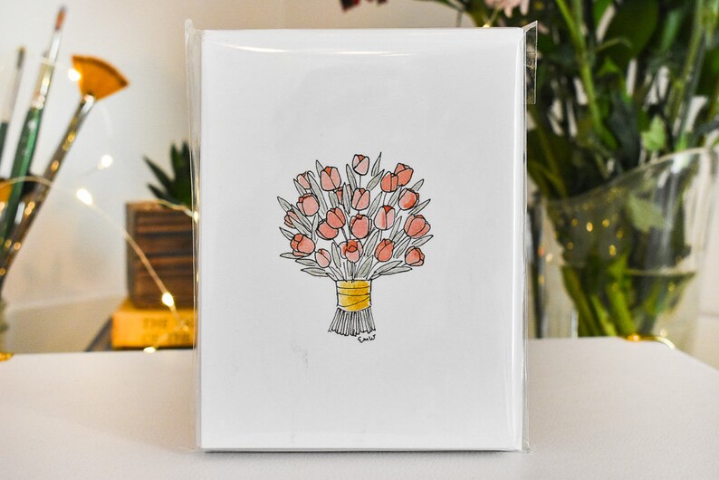 K&ouml;nnte beinhalten: Eine wei&szlig;e Gru&szlig;karte mit einer Aquarellillustration eines Bouquets aus rosa Tulpen, die mit einem gelben Band gebunden sind. Die Karte ist in einer transparenten Plastikh&uuml;lle versiegelt, mit Pinseln und Blumen im Hintergrund.