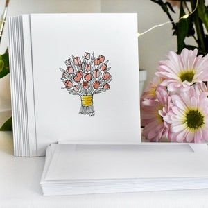 K&ouml;nnte beinhalten: Ein Stapel wei&szlig;er Gru&szlig;karten mit einer handgezeichneten Illustration eines Bouquets aus rosa Tulpen, die mit einem gelben Band zusammengebunden sind. Passende Umschl&auml;ge liegen davor. Rosa Blumen und Pinsel befinden sich im Hintergrund.