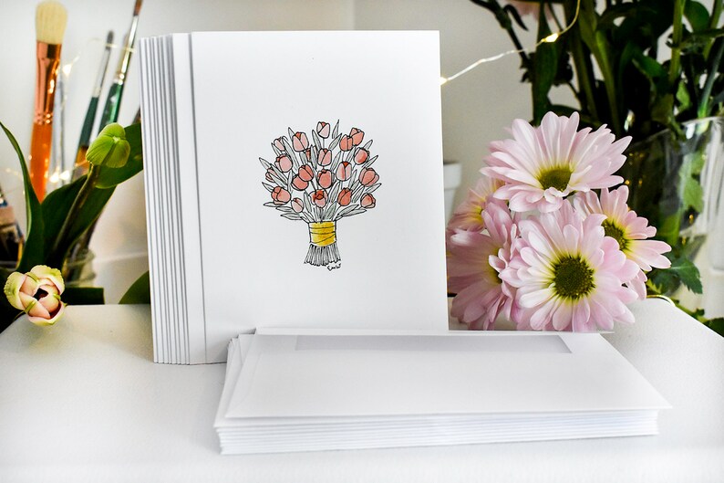 K&ouml;nnte beinhalten: Ein Stapel wei&szlig;er Gru&szlig;karten mit einer handgezeichneten Illustration eines Bouquets aus rosa Tulpen, die mit einem gelben Band zusammengebunden sind. Passende Umschl&auml;ge liegen davor. Rosa Blumen und Pinsel befinden sich im Hintergrund.