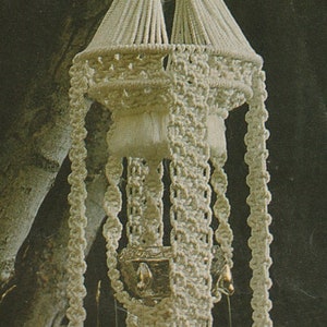May include: A white macrame plant hanger with a woven design and a metal pot. The hanger is hanging from a tree branch.