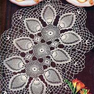 Puede incluir: Un tapete de ganchillo hecho a mano, de color crema, con un diseño floral. El tapete está colocado sobre una superficie marrón oscuro, con dos tazas de café y galletas en el fondo. Flores naranjas en la esquina inferior derecha.