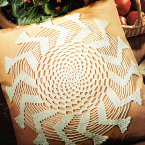 May include: A round white crochet doily featuring a complex spiral pattern with radiating zigzag spokes. The intricate needlework creates a circular design with openwork gaps between the solid stitched sections, centered on a tan fabric surface.