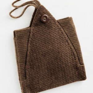 Op de afbeelding: Een donkerbruin gebreid accessoire met een driehoekige flap die wordt vastgezet met een enkele donkerbruine houten knoop. Het item heeft een gebreid lichaam met textuur, geribbelde zijpanelen en een klein gevlochten lusje aan de bovenhoek.