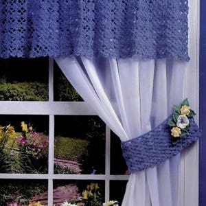 May include: A blue crocheted valance and matching tieback featuring three-dimensional fabric flowers in white, yellow, and green, paired with a sheer white curtain panel. The valance has a delicate openwork pattern and hangs from a curtain rod.