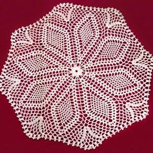 May include: A white, octagonal crocheted doily. The intricate lace pattern radiates from a central circle, with a repeating design of openwork and solid sections. The doily is set against a red background.