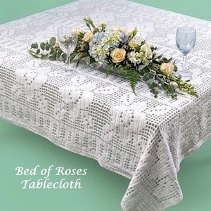 Pode incluir: Uma toalha de mesa branca, com tecido aberto e um desenho floral, colocada numa mesa quadrada. A mesa é decorada com um centro de mesa floral e dois copos de vinho. O texto "Bed of Roses Tablecloth" é visível.