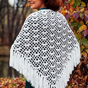 May include: A white, open-weave crochet shawl with a fringed edge. The shawl is draped over the shoulders. The background features autumn foliage with red, orange, and yellow leaves.