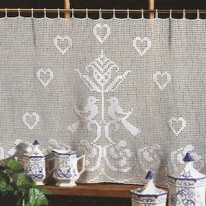 May include: White crocheted curtain with a heart and bird design. The curtain is hanging on a window with a wooden frame. There are three blue and white ceramic jars in the foreground.