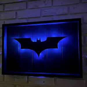 May include: A rectangular wall-mounted sign featuring a black silhouette of a bat emblem centered on a dark background. The emblem is illuminated from behind by a vibrant blue LED light, creating a glowing effect within a black frame.