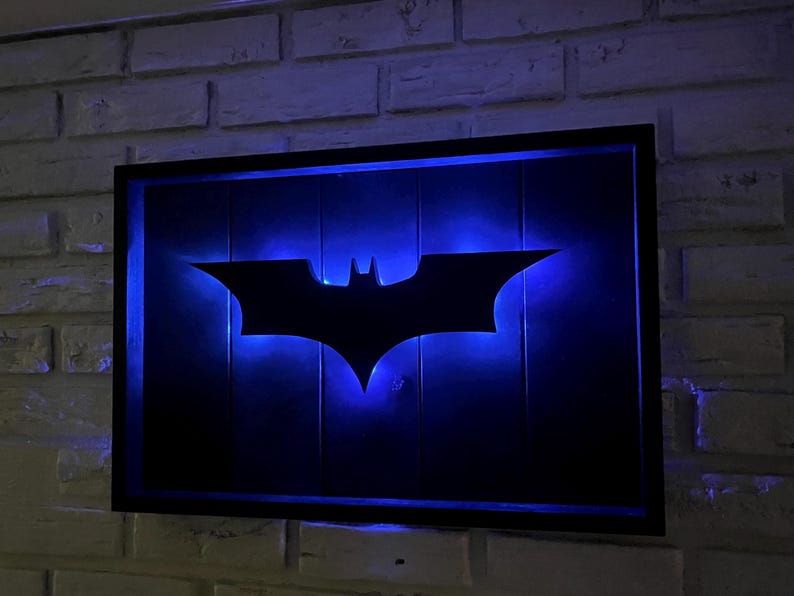 May include: A rectangular wall-mounted sign featuring a black silhouette of a bat emblem centered on a dark background. The emblem is illuminated from behind by a vibrant blue LED light, creating a glowing effect within a black frame.