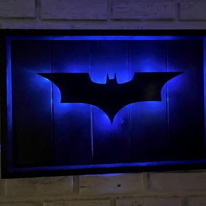 May include: A rectangular wall-mounted sign featuring a black silhouette of the Batman logo centered on a dark wooden panel. The sign is illuminated from behind by a vibrant blue LED light, creating a glowing outline around the central emblem.