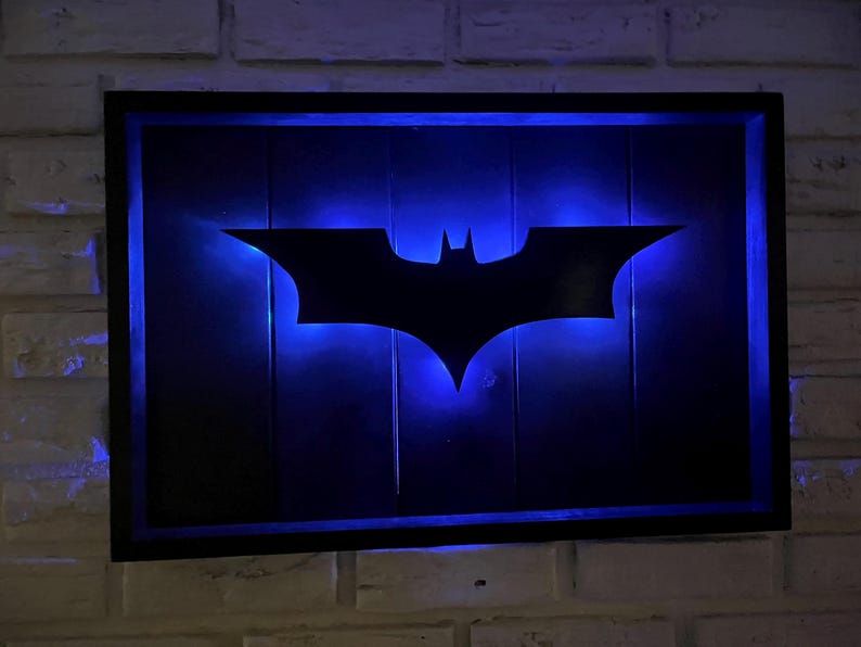 May include: A rectangular wall-mounted sign featuring a black silhouette of the Batman logo centered on a dark wooden panel. The sign is illuminated from behind by a vibrant blue LED light, creating a glowing outline around the central emblem.
