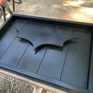 May include: A rectangular black wooden tray featuring a raised, matte black Batman bat-symbol emblem centered on the base. The tray is constructed from vertical wooden planks with a smooth, dark finish and a solid frame border surrounding the interior.